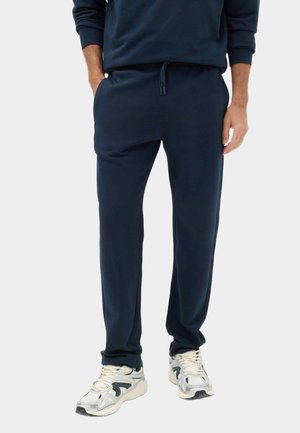 Mann trägt dunkelblaue Jogginghose mit Kordelzug in der Taille und weißen Sneakers, Hände teilweise in den Taschen, steht vor schlichtem Hintergrund.