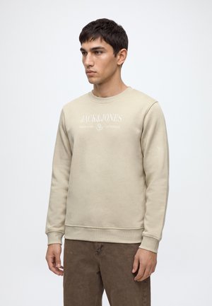 Млад мъж, носещ бежов суичър Jack & Jones и кафяви панталони, стои пред обикновен бял фон.