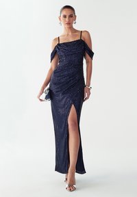 Mornarsko modra večerna obleka z bleščicami, z ramo odkritimi rokavi, stranskim razporkom in oprijetim krojem. Srebrna torbica in sandali z jermenicami v kovinskem sijaju.