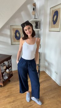 Žena sa usmieva v interiéri, oblečená v bielom tielku, námornícky modrých širokých nohaviciach, sivých teniskách a so slnečnými okuliarmi na hlave, stojaca na drevenej podlahe.