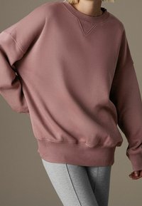 Persoon draagt een oversized sweatshirt in stoffig roze en lichtgrijze leggings, staand voor een effen gedempte bruine achtergrond.