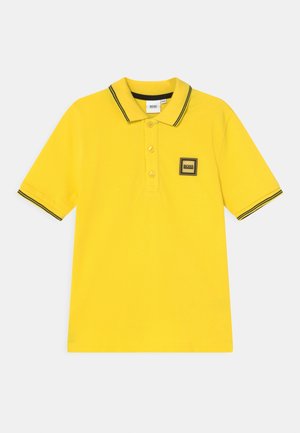 Polo giallo con colletto e polsini neri con strisce a contrasto, e un piccolo logo quadrato sul lato sinistro del petto.