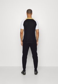 T-shirt de sport noir et blanc avec un col jaune contrasté, associé à un jogging noir et des chaussures de sport noires, vu de dos.