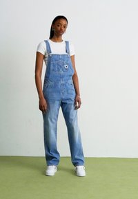 Lys blå denim overalls med justerbare stropper, en frontlomme, løs passform og falmet tekstur, paret med en tettsittende hvit t-skjorte og hvite joggesko.
