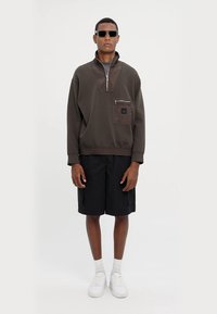 Felpa zip con cappuccio marrone scuro, dotata di tasca sul petto, abbinata a pantaloni corti neri. Il modello indossa occhiali da sole e sneakers bianche. Tessuto dalla consistenza liscia.