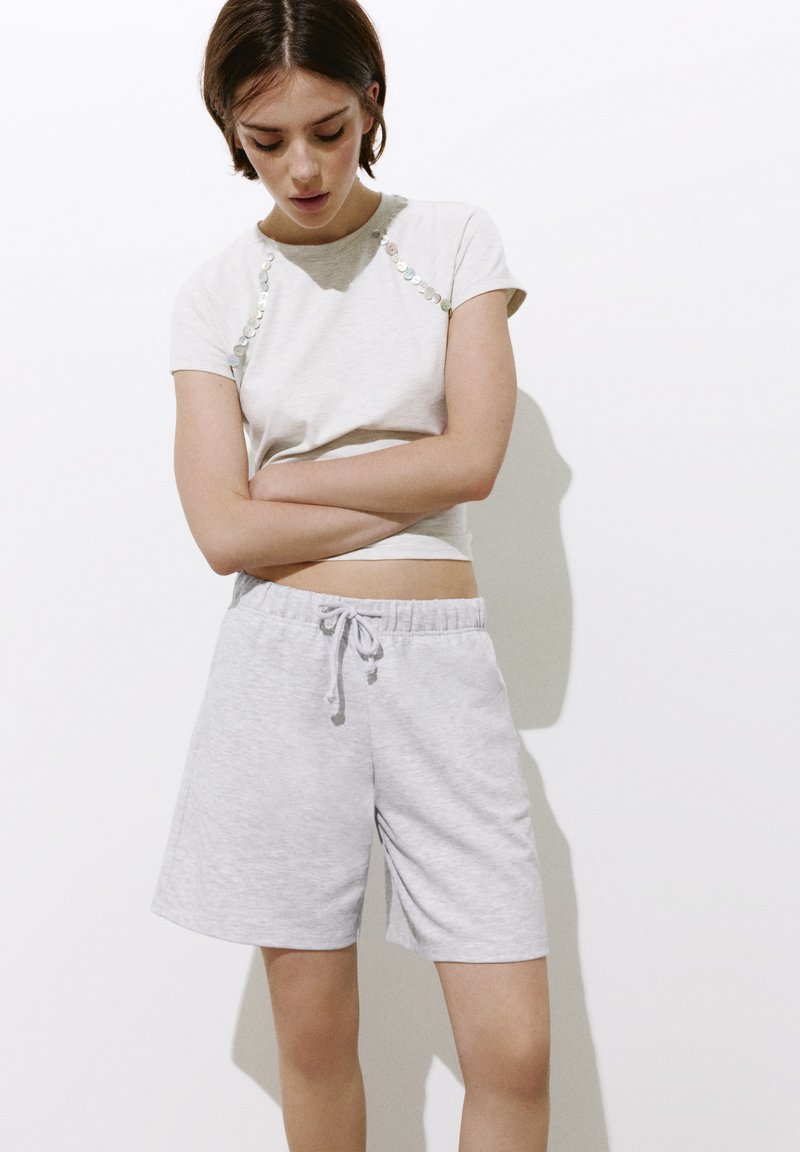 Jeune femme portant un t-shirt blanc avec des boutons et un short gris clair à cordon, debout les bras croisés et regardant vers le bas.