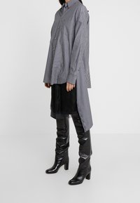 Randig lång skjorta i marinblå och vit bomull, med knäppt fram, manschetter och en rundad nedkant, tillsammans med svarta knähöga läderstövlar.