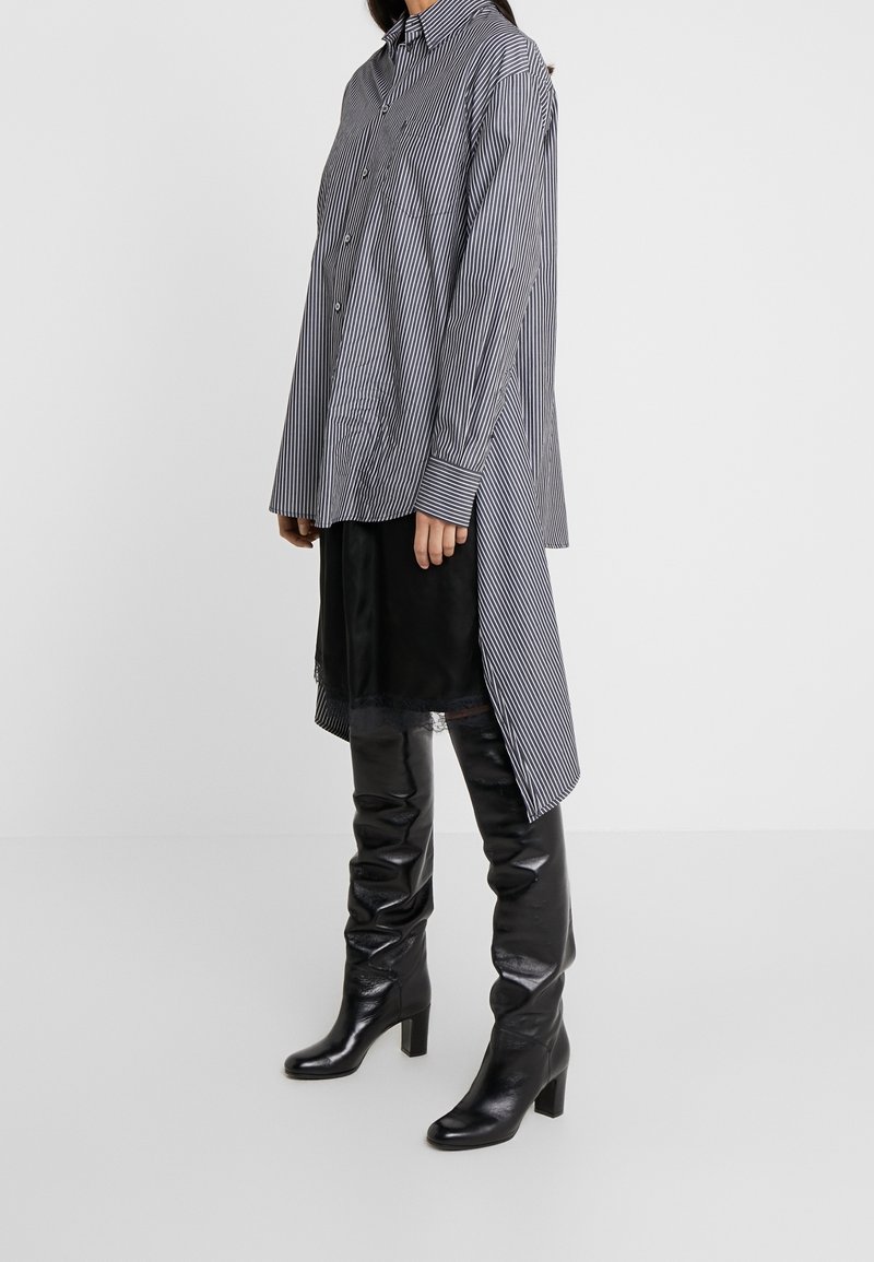 Randig lång skjorta i marinblå och vit bomull, med knäppt fram, manschetter och en rundad nedkant, tillsammans med svarta knähöga läderstövlar.