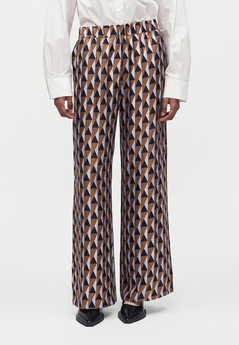 Personne portant un pantalon taille haute à jambes larges avec un motif géométrique triangulaire marron et noir, une chemise blanche à manches longues et des chaussures noires.