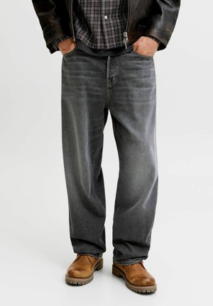 Mann trägt weite graue Jeans, braune Lederstiefel, ein kariertes Hemd und eine dunkle Lederjacke mit den Händen in den Taschen.