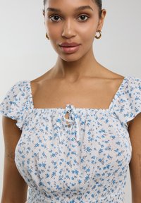 Vestido de tela ligera blanca con un estampado floral azul. Presenta mangas cortas con volantes y un detalle de lazo en el escote.