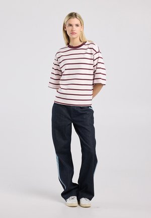 Jeune femme debout, les mains derrière le dos, portant une chemise rayée blanche et bordeaux, un jean large foncé avec des panneaux latéraux clairs et des baskets blanches.