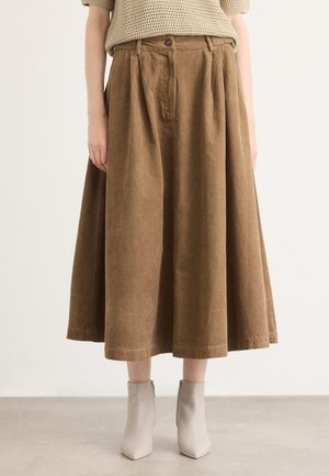 Jupe midi en velours côtelé marron à plis associée à des bottines beiges portée par une personne habillée d'un haut en maille beige, debout sur un sol clair contre un mur uni.