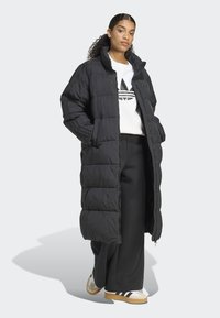 Γυναίκα που φοράει ένα μακρύ μαύρο καπιτονέ παλτό, λευκή μπλούζα με μαύρο λογότυπο, μαύρο παντελόνι φαρδύ και λευκά αθλητικά παπούτσια με μαύρες ρίγες.