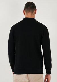 Pulóver negro de manga larga con textura acanalada, cuello y puños ajustados. El dobladillo es ligeramente elástico, diseñado para un ajuste relajado.
