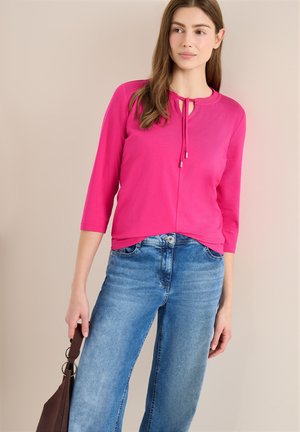 Donna che indossa una maglietta rosa acceso a maniche 3/4 con fiocco al collo e jeans blu slavati, che tiene una borsa marrone scuro su sfondo beige.