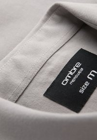 Gros plan sur le col d'une chemise en tissu gris avec une étiquette noire cousue à l'intérieur indiquant "Oxbore menswear taille M".