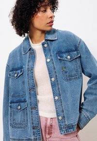 Lichtblauwe denimjack met zilveren knopen, twee borstzakken en oranje stiksels. Draagt over een crèmekleurige trui met een zachte textuur.