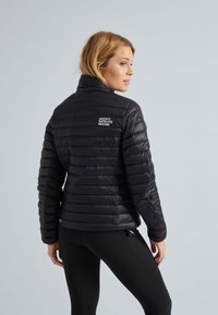 Doudoune noire avec texture matelassée, col haut et détail de logo au dos. Présente un design ajusté et est portée par-dessus des leggings noirs.