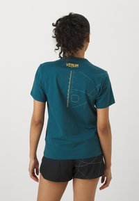 Teal puuvillane t-särk lühikeste varrukatega, millel on kollane "VENUM" logo ja abstraktsed geomeetrilised mustrid seljal.
