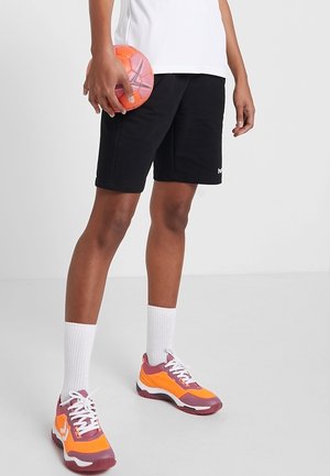 Personne debout portant un short noir, des chaussettes blanches, des baskets orange et rose, tenant un ballon de handball orange et rose dans sa main droite.