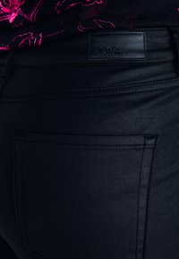 Jean en denim noir avec une texture lisse, doté de coutures sur les poches arrière et d'une étiquette de marque en cuir rectangulaire à la taille.