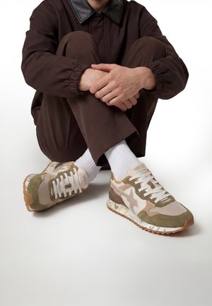 Person in braunem Outfit sitzt im Schneidersitz und trägt beige und olivgrüne Sneakers mit weißen Schnürsenkeln und weißen Socken auf weißem Hintergrund.