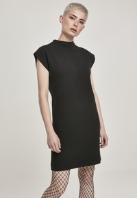 Zwarte mouwloze jurk met een hoge hals, ribbelstructuur en een rechte silhouet, gecombineerd met visnetkousen.