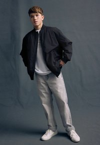 Svart bomberjacka med dragkedja, vit grafisk t-shirt under, grå baggy jeans och vita sneakers. Enkel design, neutrala färger, avslappnad passform.