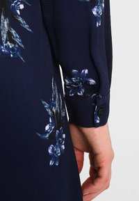 Abito navy con stampe floreali in blu e bianco, caratterizzato da un tessuto testurizzato e un dettaglio con bottoni sui polsini delle maniche lunghe.