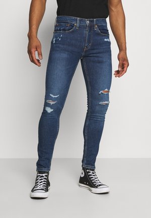 Homme portant un jean skinny bleu délavé et des baskets montantes noires, debout sur un fond blanc.