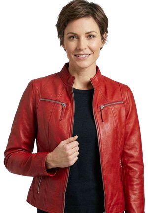 Woman with short brown hair wearing a red leather jacket over a black top, smiling and holding the jacket zipper near the collar.