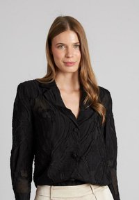 Zwarte blouse met een textuur, een bloemenpatroon, knoopsluiting en een geknipt colbert. Lange mouwen en lichte stof.