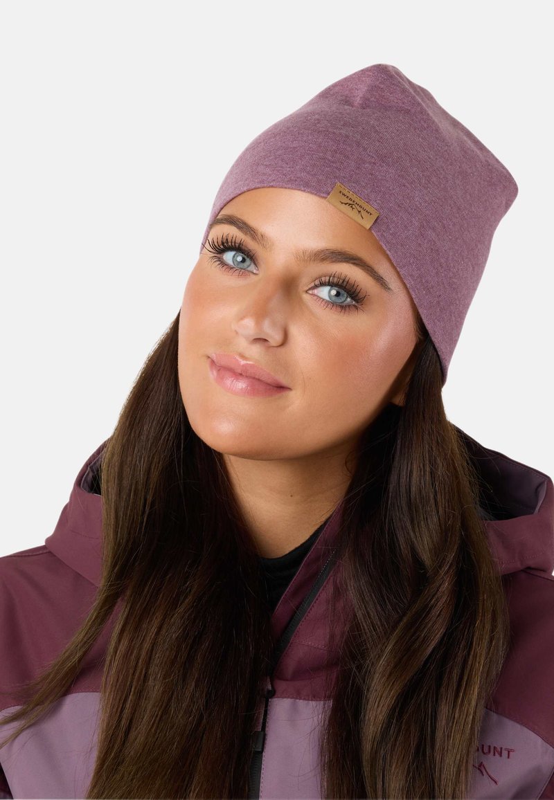 Lila beanie muts gemaakt van zacht materiaal, met een iets slouchy ontwerp, voorzien van een klein bruin labelaccent aan de voorkant, gedragen met een paarse jas.