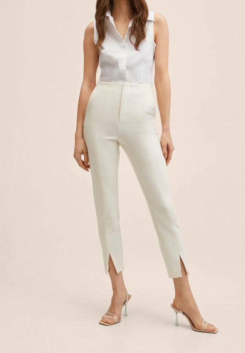 Femme portant un chemisier blanc sans manches, un pantalon crème taille haute avec des fentes à l'avant aux chevilles, et des sandales à talons hauts à brides transparentes.