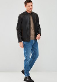 Chaqueta de cuero negra con acentos acolchados en los hombros, cierre frontal y bolsillos con cremallera. Llevada sobre una camisa beige y combinada con jeans azules y botas negras.