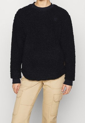 Osoba ubrana w czarny teksturowany polarowy sweter i beżowe spodnie typu cargo z kieszeniami, stojąca na jednolitym jasnym tle.