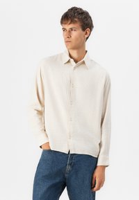 Beige long-sleeve shirt with a textured weave, button-down collar, and front placket. Paired with blue denim jeans.
