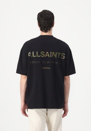 Mężczyzna noszący czarny oversize T-shirt z tekstem "ALLSAINTS" w kamuflażu, japońskimi znakami i napisem "LONDON" na plecach, w połączeniu z jasnymi spodniami.
