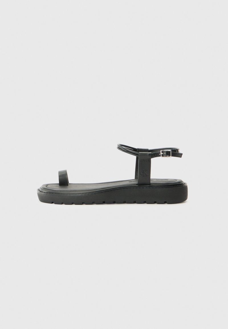 Sandalha preta plana com uma sola de borracha texturizada. Apresenta uma única tira para os dedos e uma tira ajustável para o tornozelo. Design minimalista com linhas limpas.