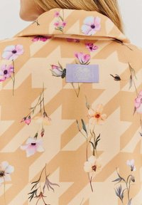 Béžová látka s květinovými vzory v růžové, fialové a žluté barvě, s designem ve tvaru cik-cak. Obsahuje štítek s nápisem "Butter Toast."