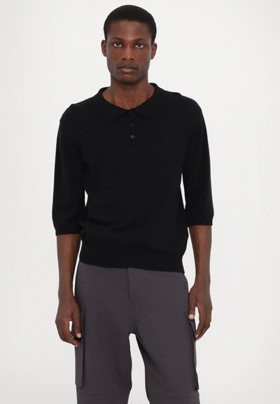 Zwart korter poloshirt met een platte kraag, drie knopen en een ribgebreide zoom. Soepele textuur. Draagt met grijze cargobroek.