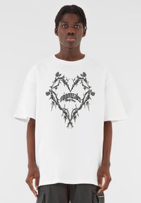 Hvit oversized t-skjorte med et svart tornerikt hjerte-design med stilisert tekst i midten. Korte ermer og løs passform.