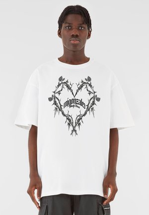 Hvit oversized t-skjorte med et svart tornerikt hjerte-design med stilisert tekst i midten. Korte ermer og løs passform.
