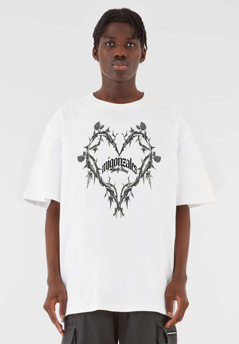 Hvit oversized t-skjorte med et svart tornerikt hjerte-design med stilisert tekst i midten. Korte ermer og løs passform.