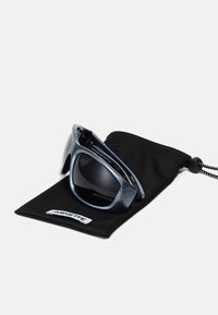 Occhiali da sole con montatura lucida di colore blu scuro, lenti grigie e un design sportivo, posizionati su un sacchetto di stoffa nera etichettato 'ARNETTE'.