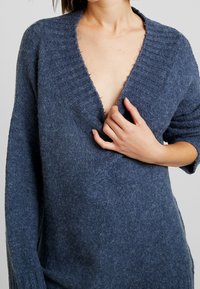 Maglione blu in lana con scollo a V profondo, collo a coste e vestibilità ampia. La texture appare morbida e leggermente pelosa. Maniche lunghe ben in evidenza.
