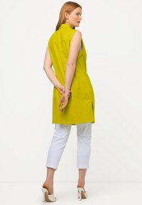 Túnica sin mangas de un brillante color amarillo con cuello y aberturas laterales, combinada con pantalones blancos hasta el tobillo y zapatos de tacón alto beige.