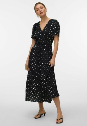 Kvinde iført en sort midikjole med hvide polkaprikker og puffede ærmer, kombineret med sorte sandaler med lave hæle på en ensfarvet hvid baggrund.