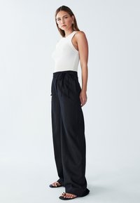 Ženska v beli brezrokavni rebrasti majici, visokih črnih širokih hlačah z zavezanimi pasom in črnih sandalih z odprtimi prsti na enostavni podlagi.